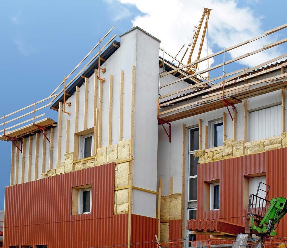 Maison en construction avec bardage blanc et rouge, charpente apparente et échafaudages sur fond de ciel bleu