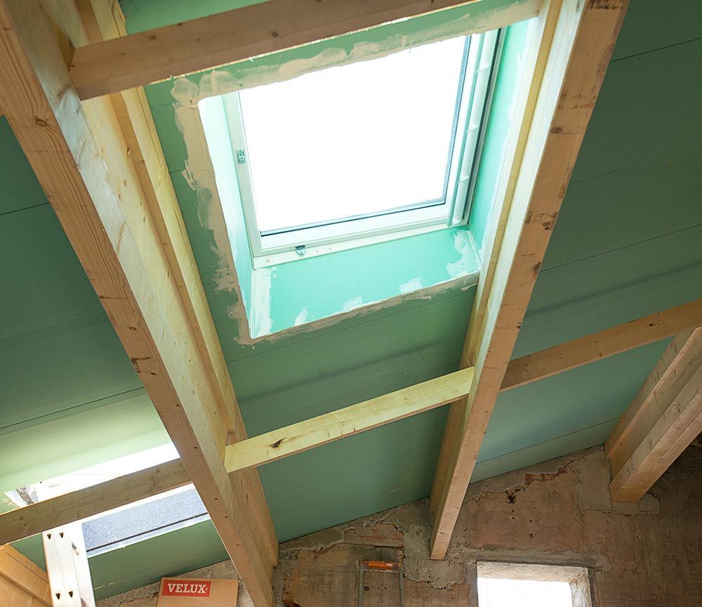 Toiture du grenier avec poutres en bois apparentes et puits de lumière, laissant apparaître des panneaux de plafond verts non finis.