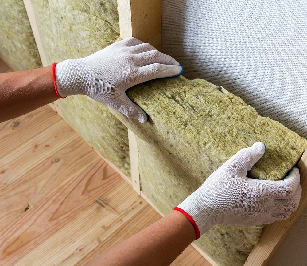 Des mains gantées de blanc ajustent un panneau isolant en fibre de verre jaune dans une ossature murale en bois.