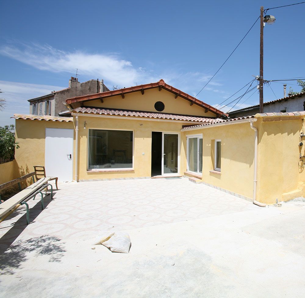 Cour ensoleillée avec maison en stuc jaune, grandes fenêtres et porte-fenêtre coulissante.