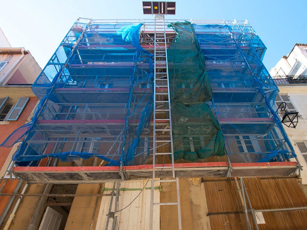 Bâtiment recouvert de filets d'échafaudage bleus et verts, avec une échelle au centre.