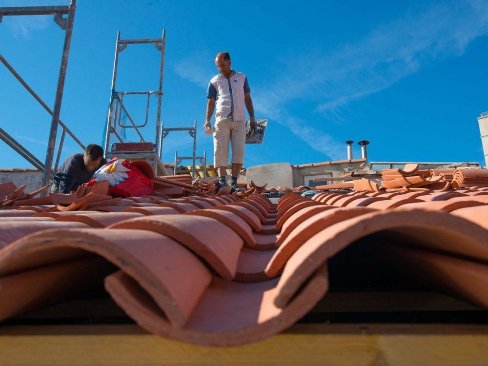 Des ouvriers travaillent sur un toit de tuiles en terre cuite sous un ciel bleu, l'un d'eux se tenant près d'un échafaudage.