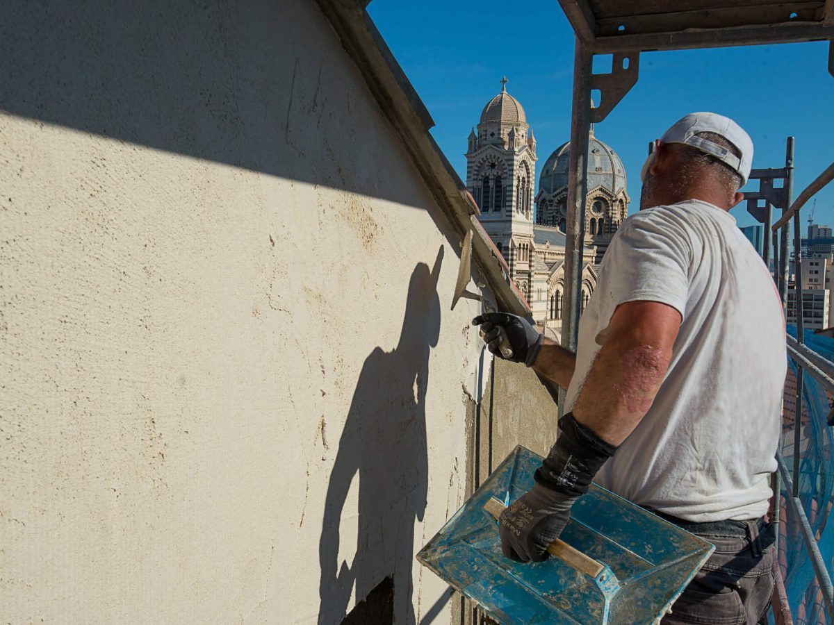 Un ouvrier peint un mur sur un échafaudage, tenant un plateau bleu avec une cathédrale en arrière-plan.