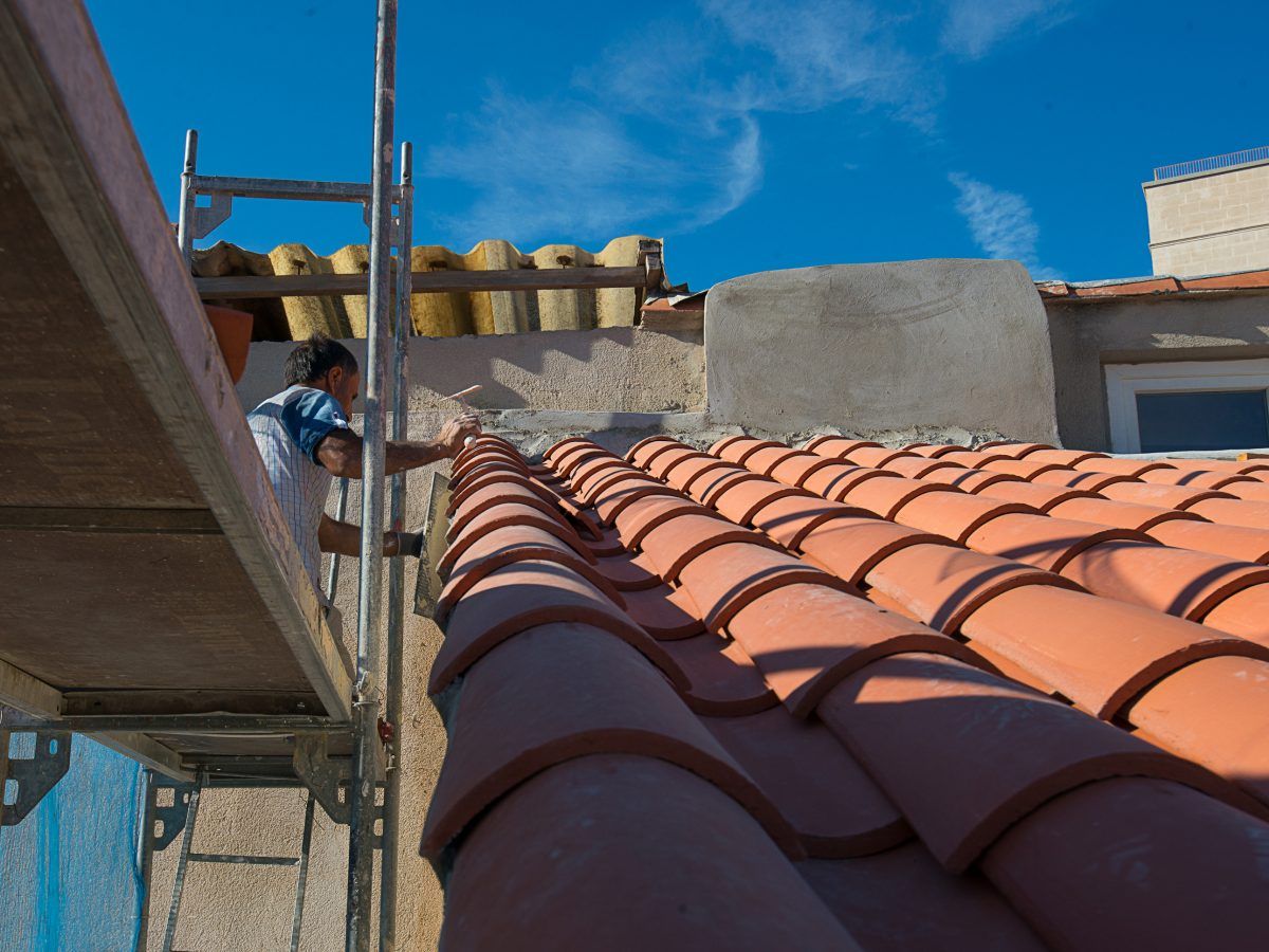 Ouvrier sur un échafaudage à côté d'un toit de tuiles rouges sous un ciel bleu, avec des bâtiments en arrière-plan