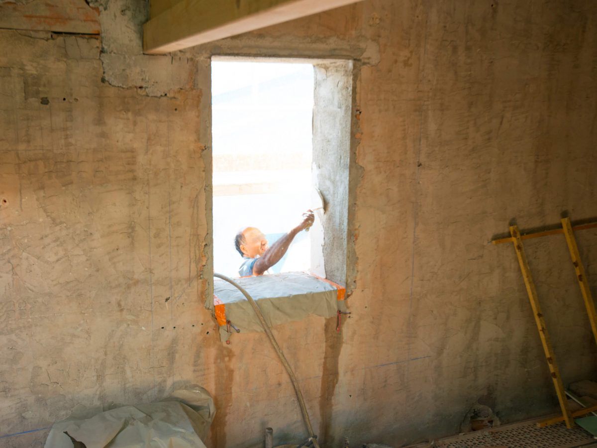 Ouvrier en train de peindre une ouverture de fenêtre sur un mur en béton brut à l'intérieur d'une pièce inachevée