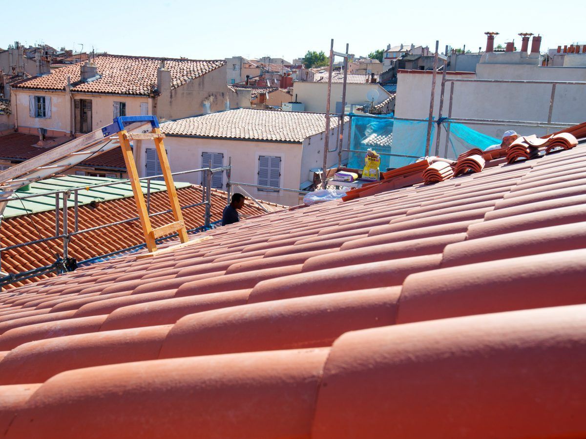 Toiture avec des tuiles rouges, des échelles et un ouvrier surplombant un quartier sous un ciel dégagé