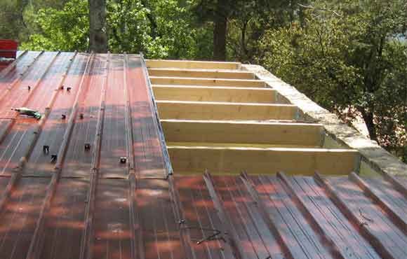 Charpente de toit en bois partiellement installée à côté d'un toit en métal rouge dans une zone boisée