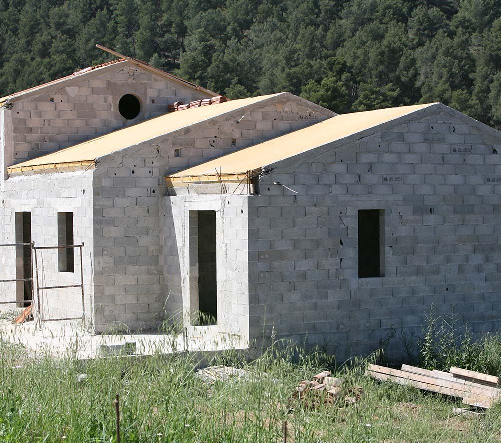Maisons en parpaings de béton inachevées, aux toits beiges, au milieu d'un champ herbeux, bordées d'arbres et de collines.