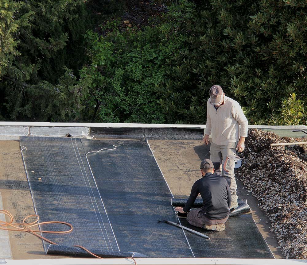 Deux ouvriers installent une membrane d'étanchéité noire sur un toit plat, avec des outils et des débris à proximité.