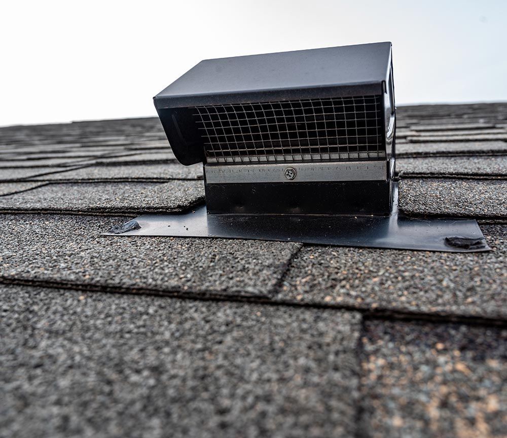 Grille d'aération noire sur un toit en bardeaux gris, se détachant sur un ciel pâle.