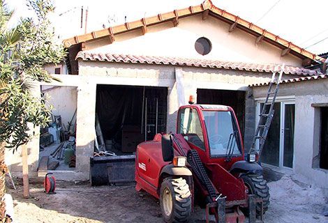 Un tracteur-chargeur rouge se trouve devant une maison en rénovation, avec des débris de construction et un garage ouvert.