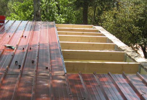Toiture en bois partiellement construite avec charpente apparente, jouxtant un toit en métal rouge, entourée d'arbres