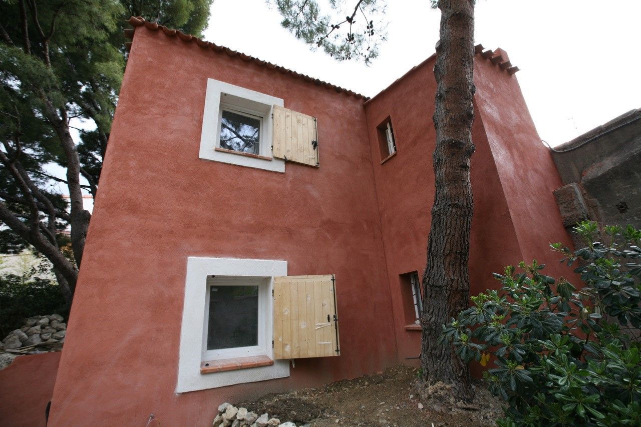 Petite maison en stuc rouge avec des fenêtres à encadrement blanc dans une zone boisée