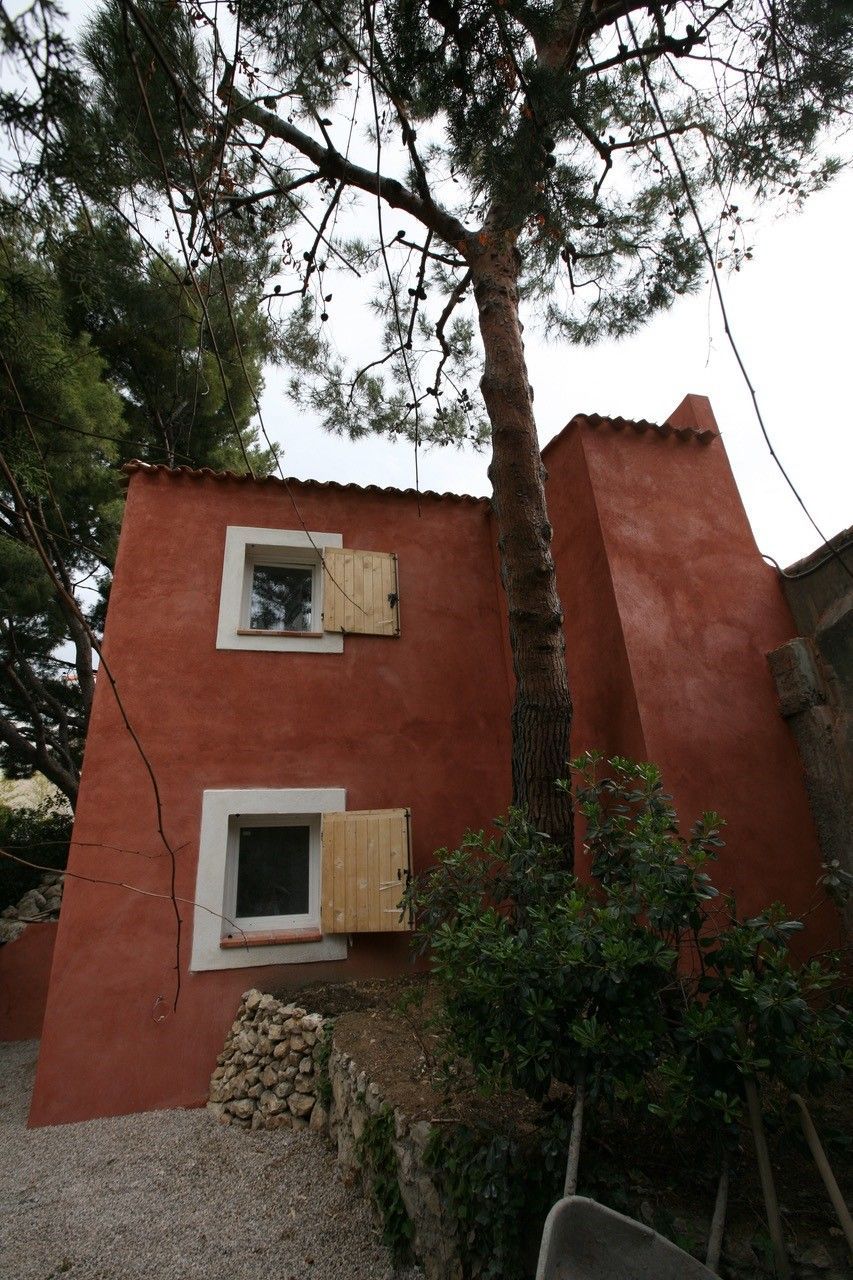 Maison en stuc rouge avec deux fenêtres à côté d'un grand arbre et de verdure