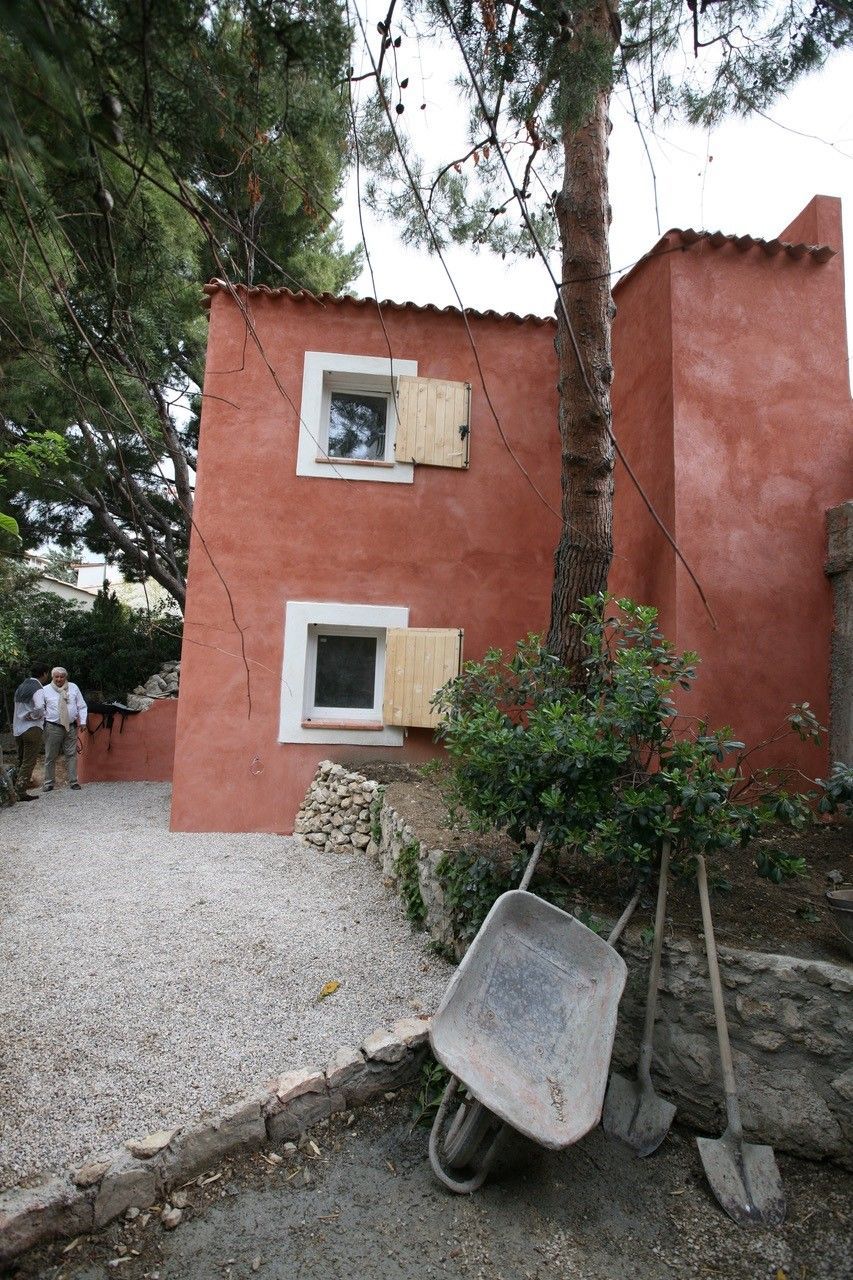 Bâtiment rouge-orange avec deux fenêtres, près d'arbres et d'une brouette sur du gravier.