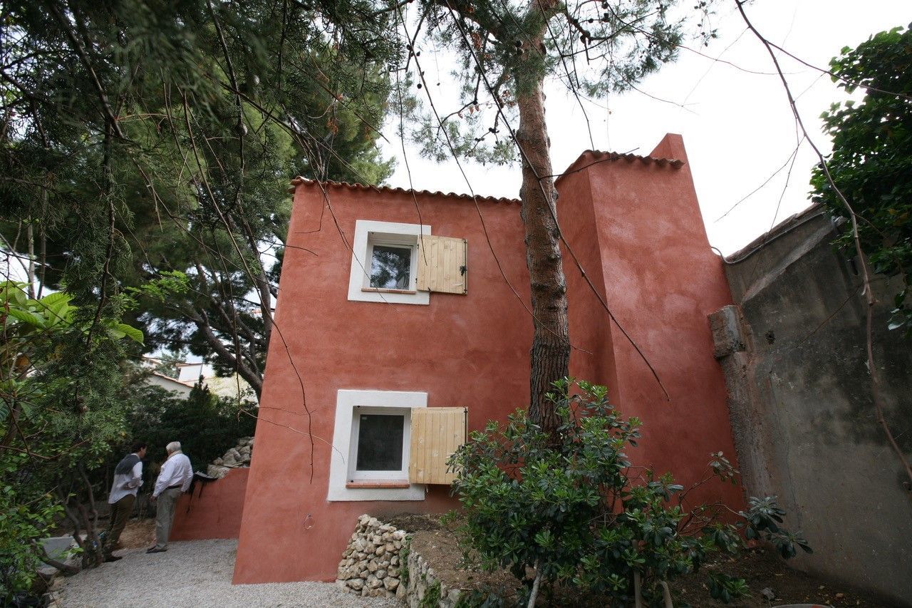 Petite maison rouge aux fenêtres à encadrement blanc, située à côté d'un grand arbre et d'un mur de pierre dans une zone boisée.