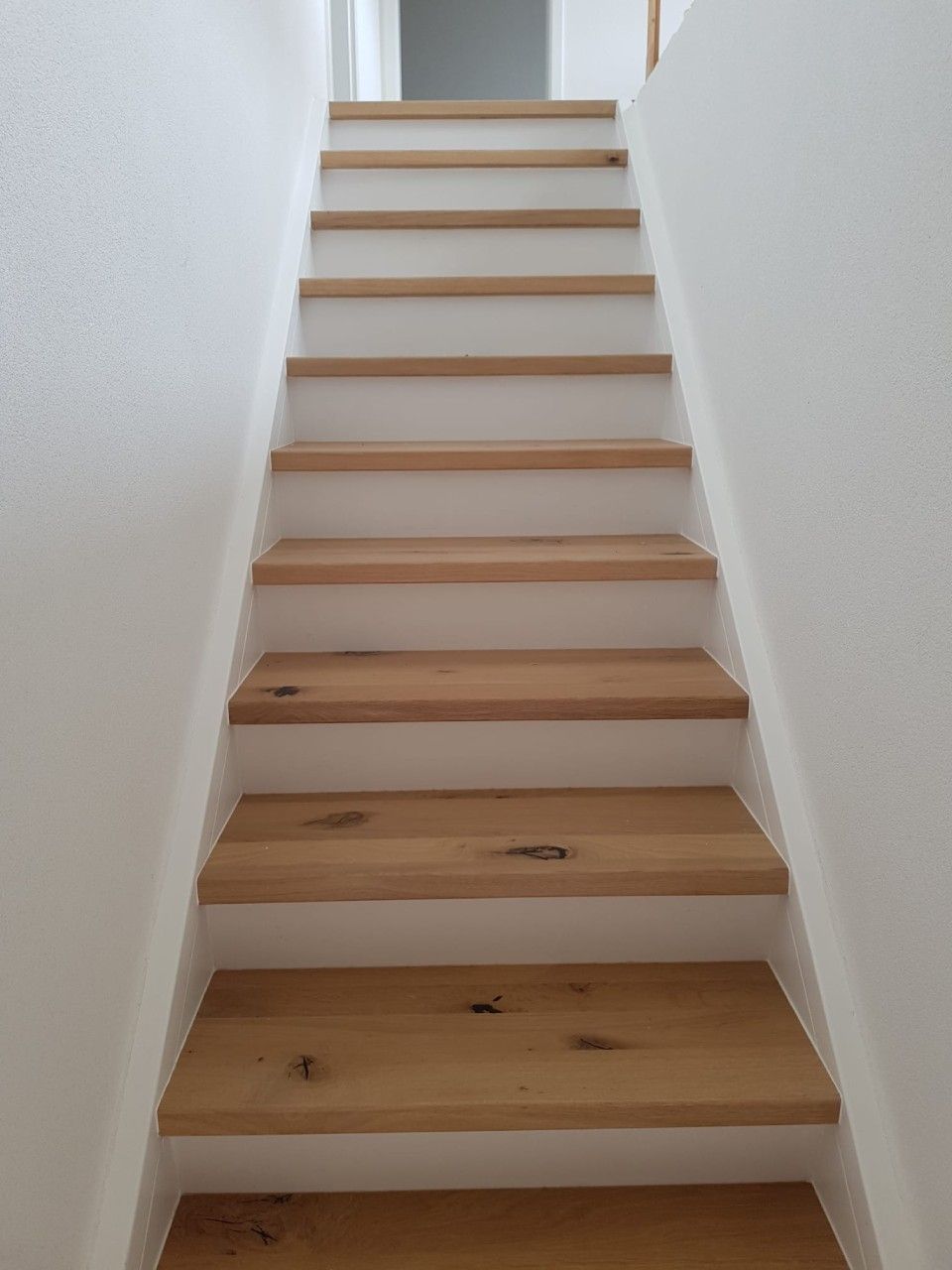 Holztreppe mit hellen Stufen, weißen Wänden, die nach oben führt.
