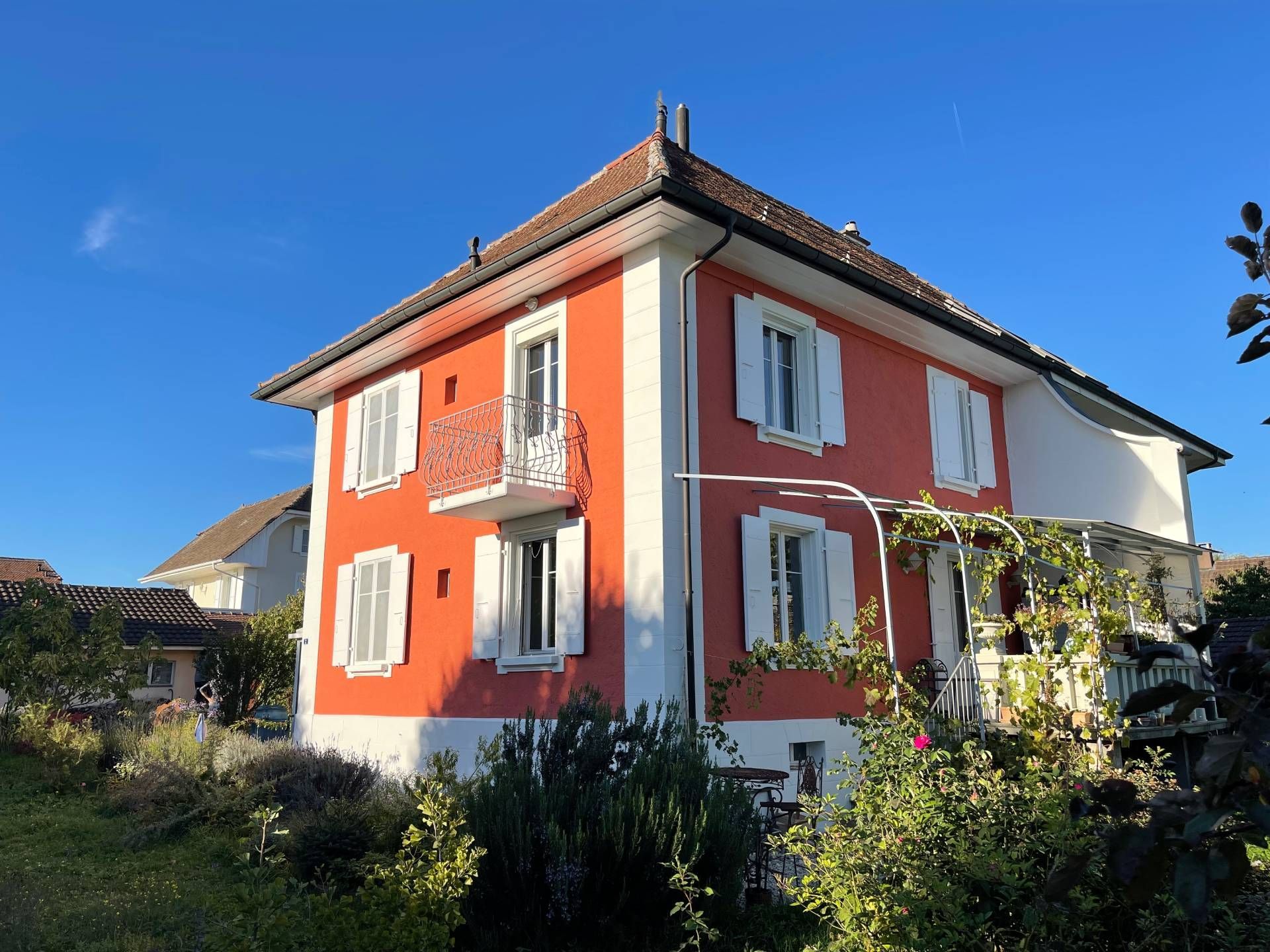 Maison orange à deux étages avec volets et boiseries blanches ; ciel bleu et verdure au premier plan.