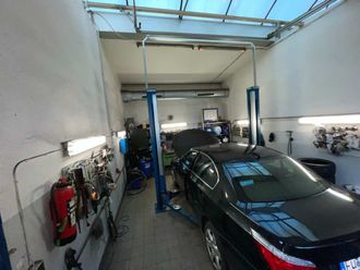 Ein schwarzer Wagen steht in einer kleinen Autowerkstatt mit Hebebühne, Werkzeug an der Wand und einem weiteren Auto im Hintergrund.