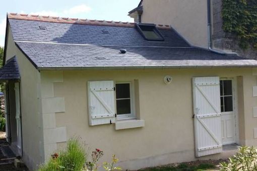 Petite maison couleur crème avec un toit en bardeaux gris et des volets et portes blancs.