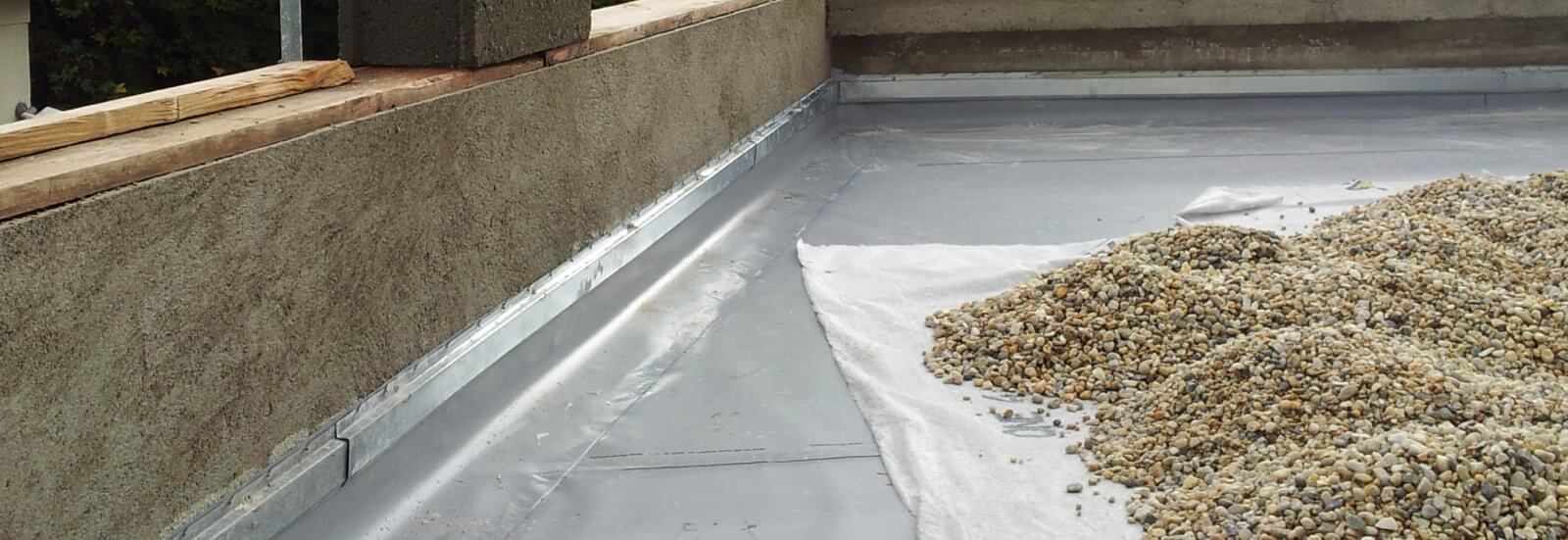 Toit plat recouvert d'une membrane d'étanchéité grise et d'un tas de gravier à côté d'un muret en briques.