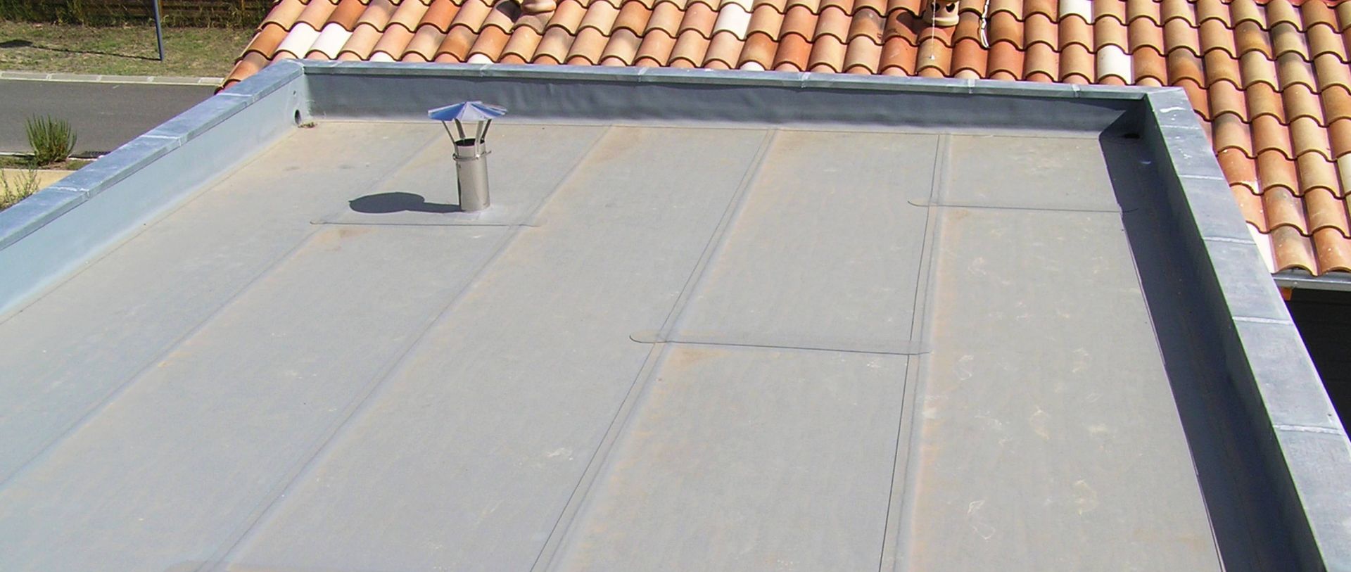 Toit plat gris avec un tuyau de ventilation et des tuiles en terre cuite visibles sur les bords