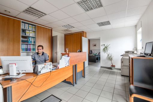 Intérieur de bureau avec une personne à un bureau, des meubles en bois, des ordinateurs et un sol carrelé clair.