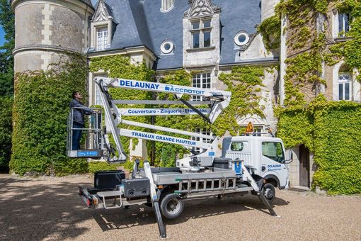 Un camion utilitaire blanc à nacelle repliée est stationné à côté d'un bâtiment en pierre recouvert de lierre.