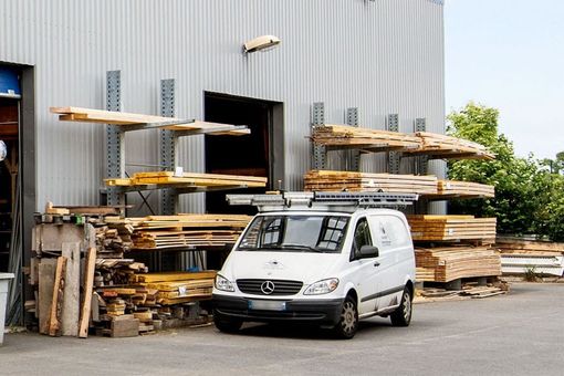 Une camionnette blanche est garée à côté de bois empilé devant un entrepôt industriel.