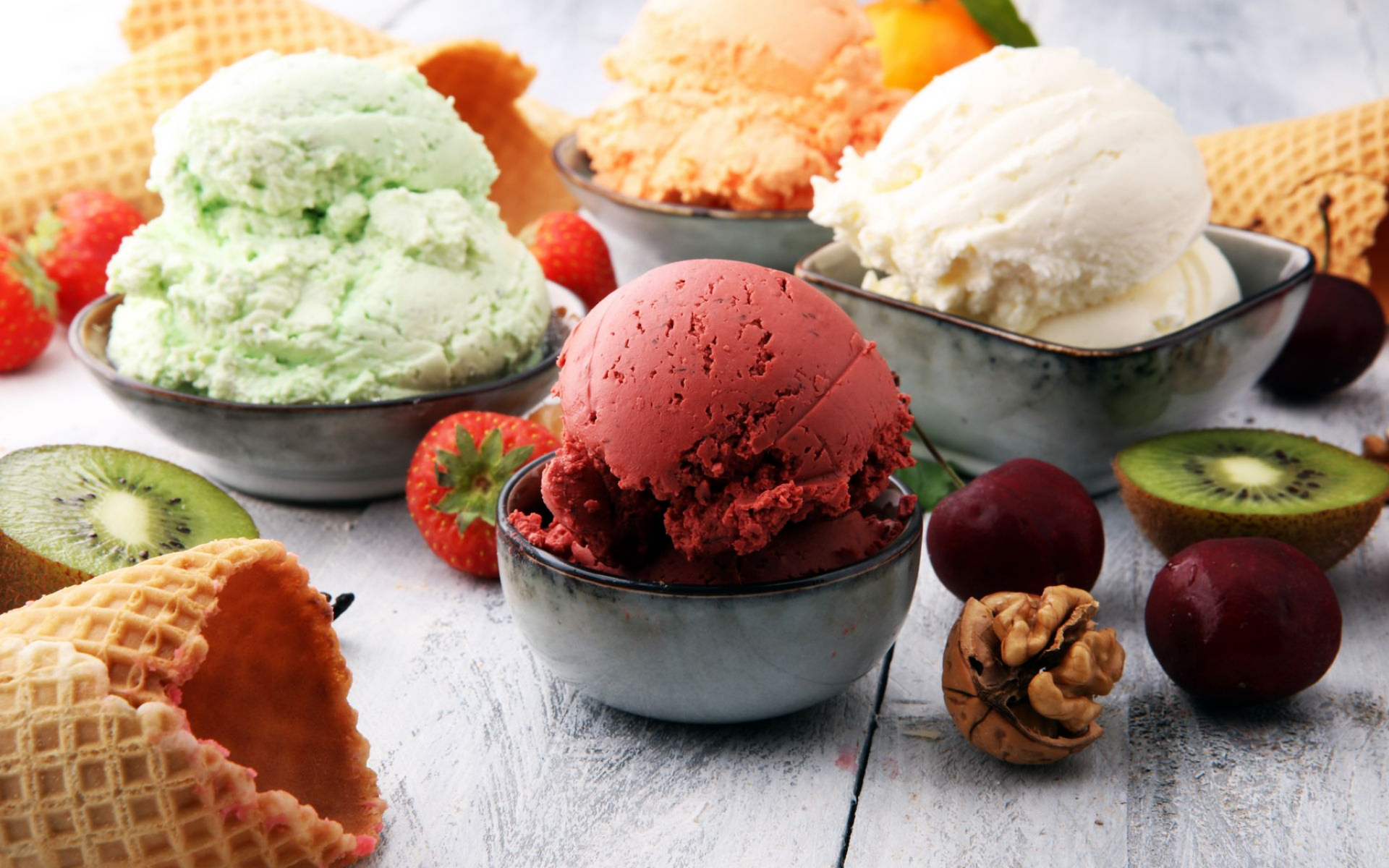 Diverses boules de glace colorées présentées dans des bols et accompagnées de cornets gaufrés et de fruits.