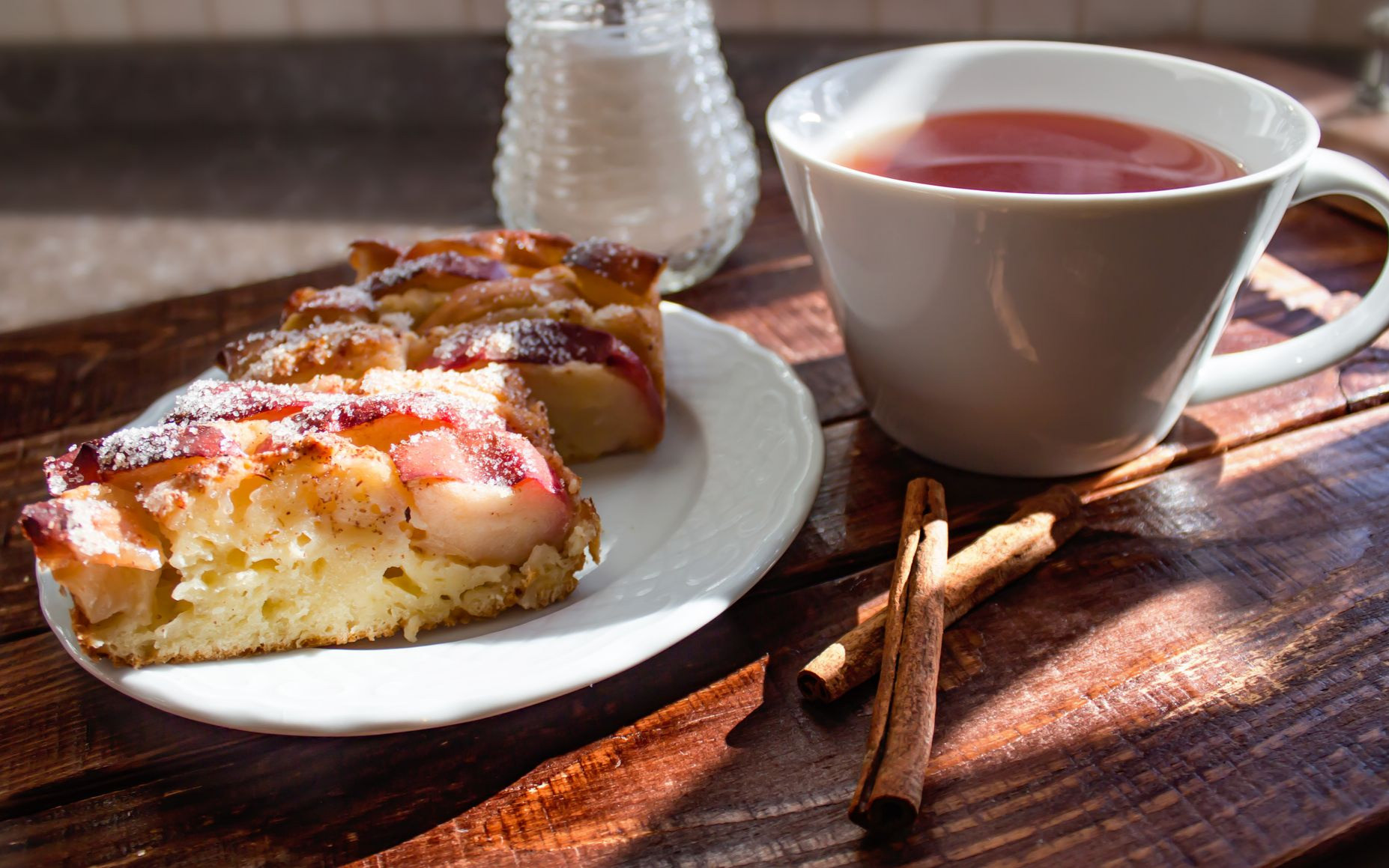Des parts de gâteau sur une assiette, une tasse de thé, du sucre et des bâtons de cannelle sur une table en bois.