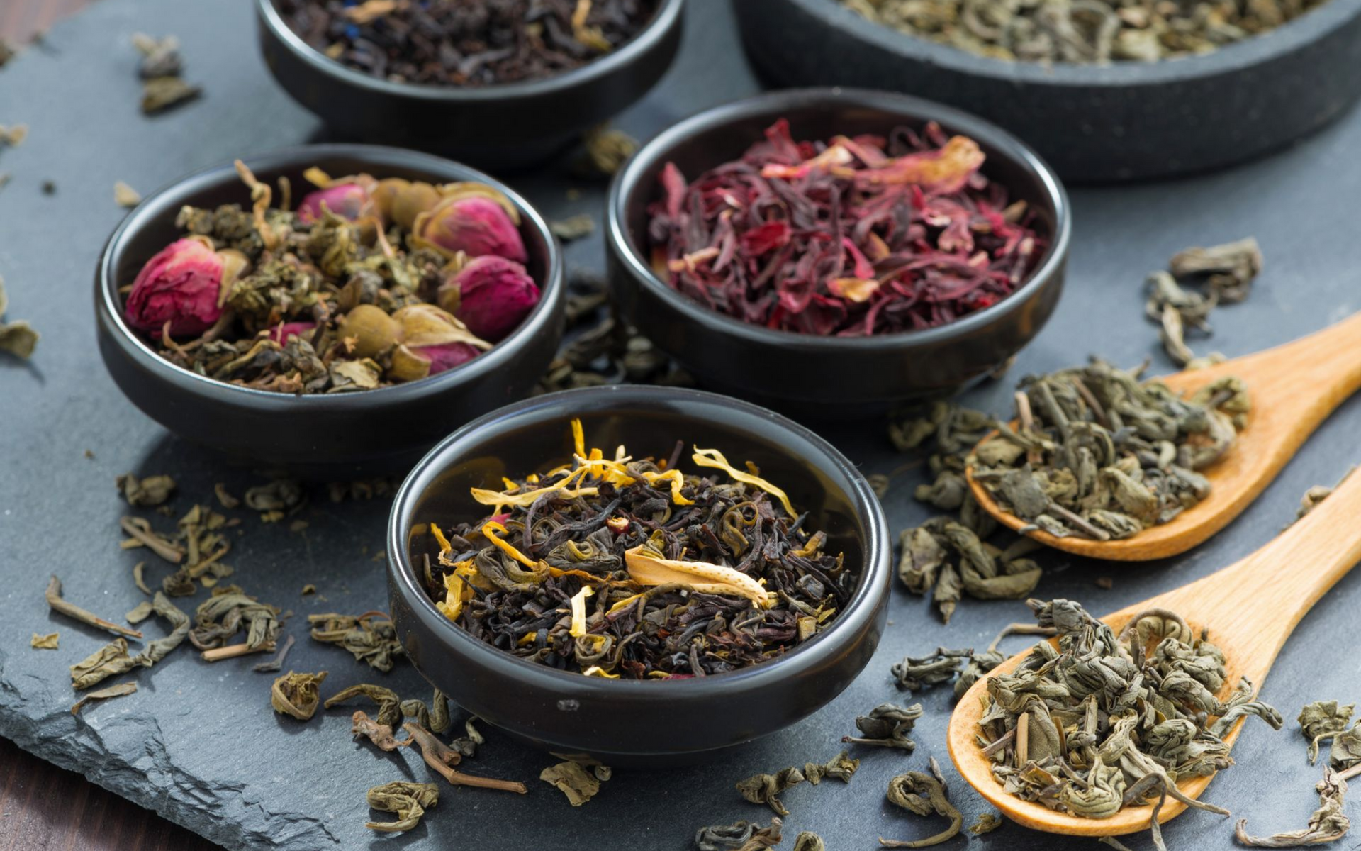 Bols de thé en vrac variés avec des cuillères en bois sur une ardoise.