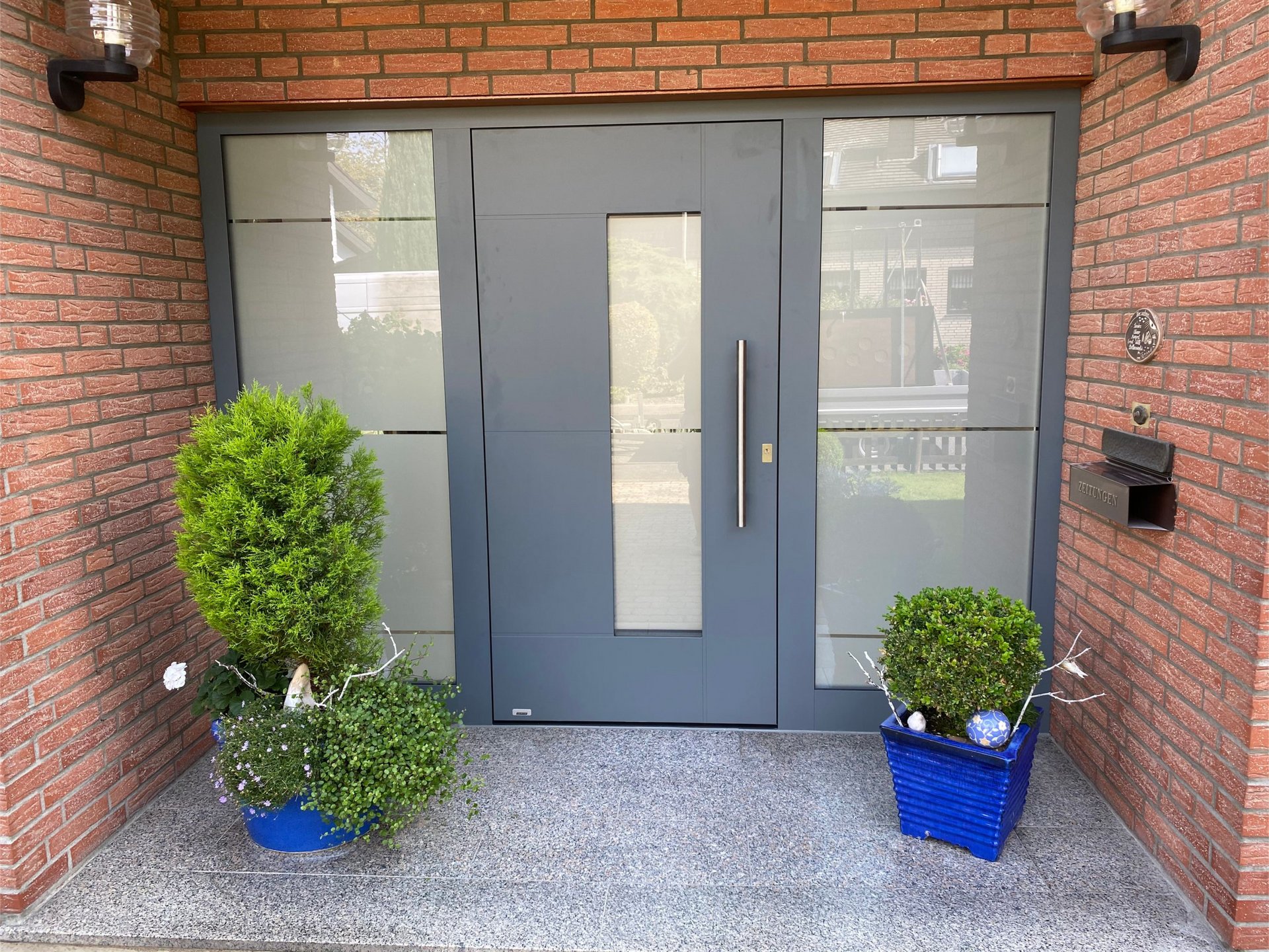 Eine moderne graue Eingangstür mit einer vertikalen Glasscheibe, flankiert von seitlichen Milchglasscheiben.