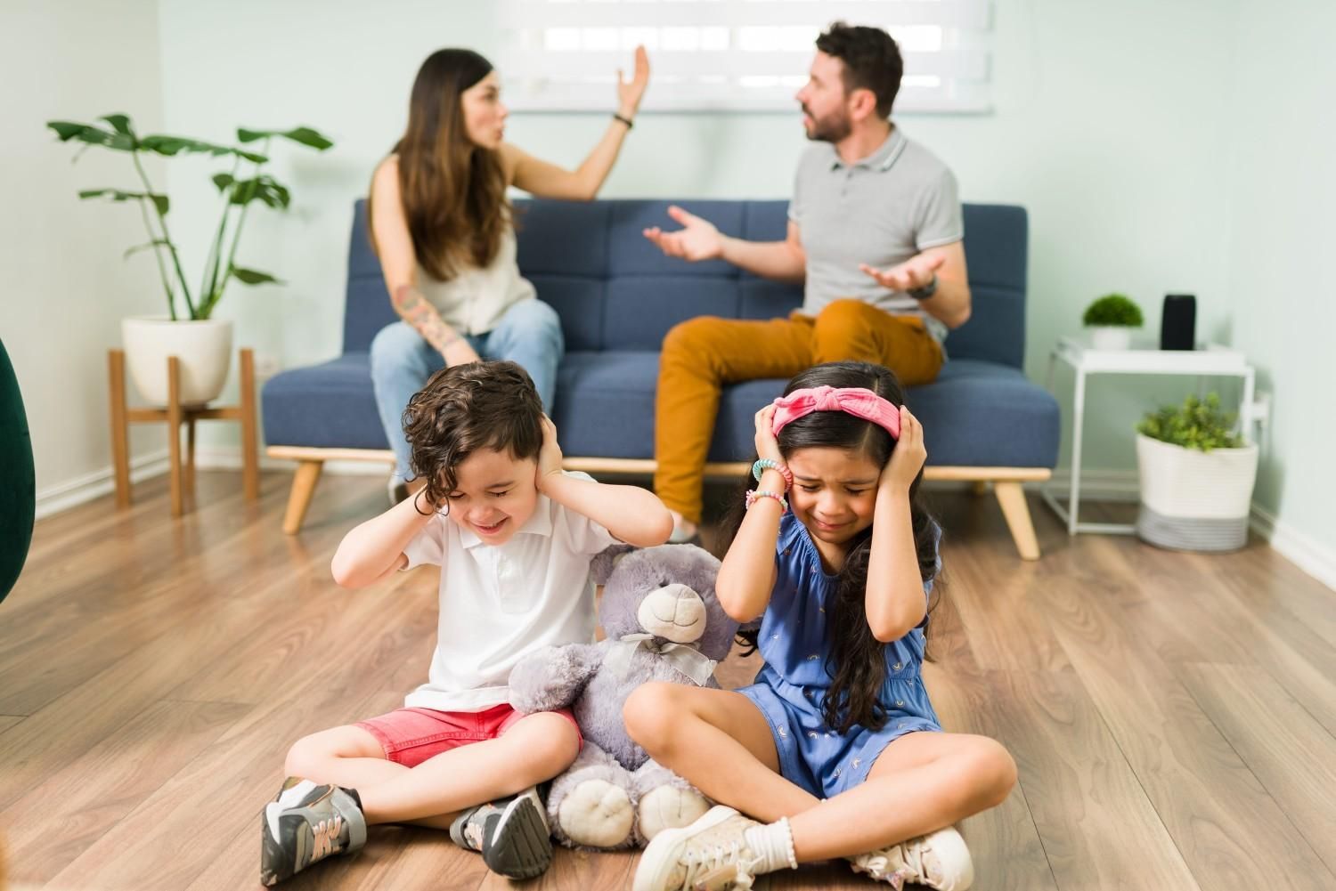 Dos niños están sentados en el suelo, tapándose los oídos, mientras dos adultos discuten en el sofá al fondo.