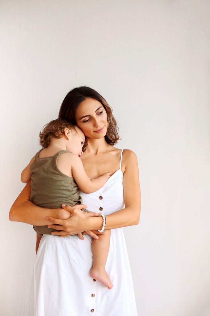 Una persona vestida con un vestido blanco abotonado sostiene a un niño que lleva un pelele verde.