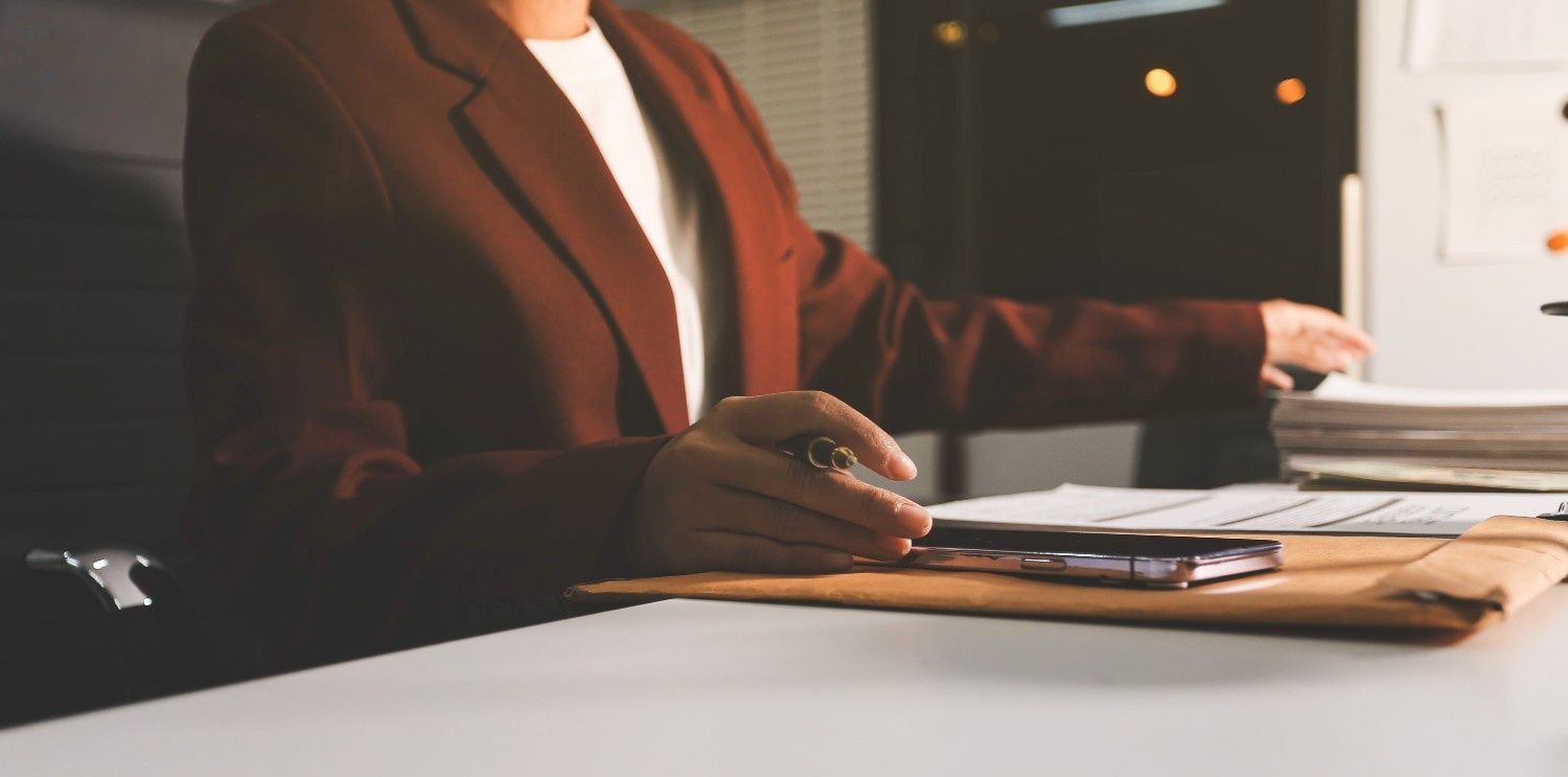 Una persona con un blazer marrón está sentada en un escritorio, con la mano apoyada sobre una pila de papeles.