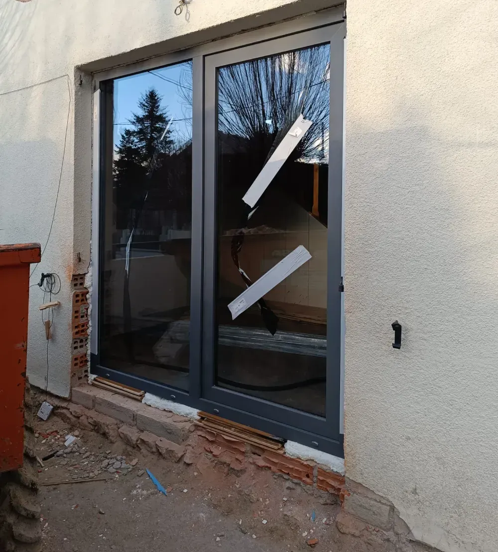 Porta corredissa de vidre amb marc fosc en una paret d'estuc, parcialment coberta amb cinta blanca, en una obra