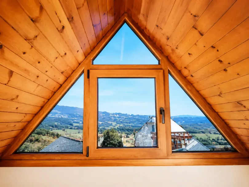 Ventana triangular de madera en el ático que enmarca una vista de montañas de un azul brillante y tejados.