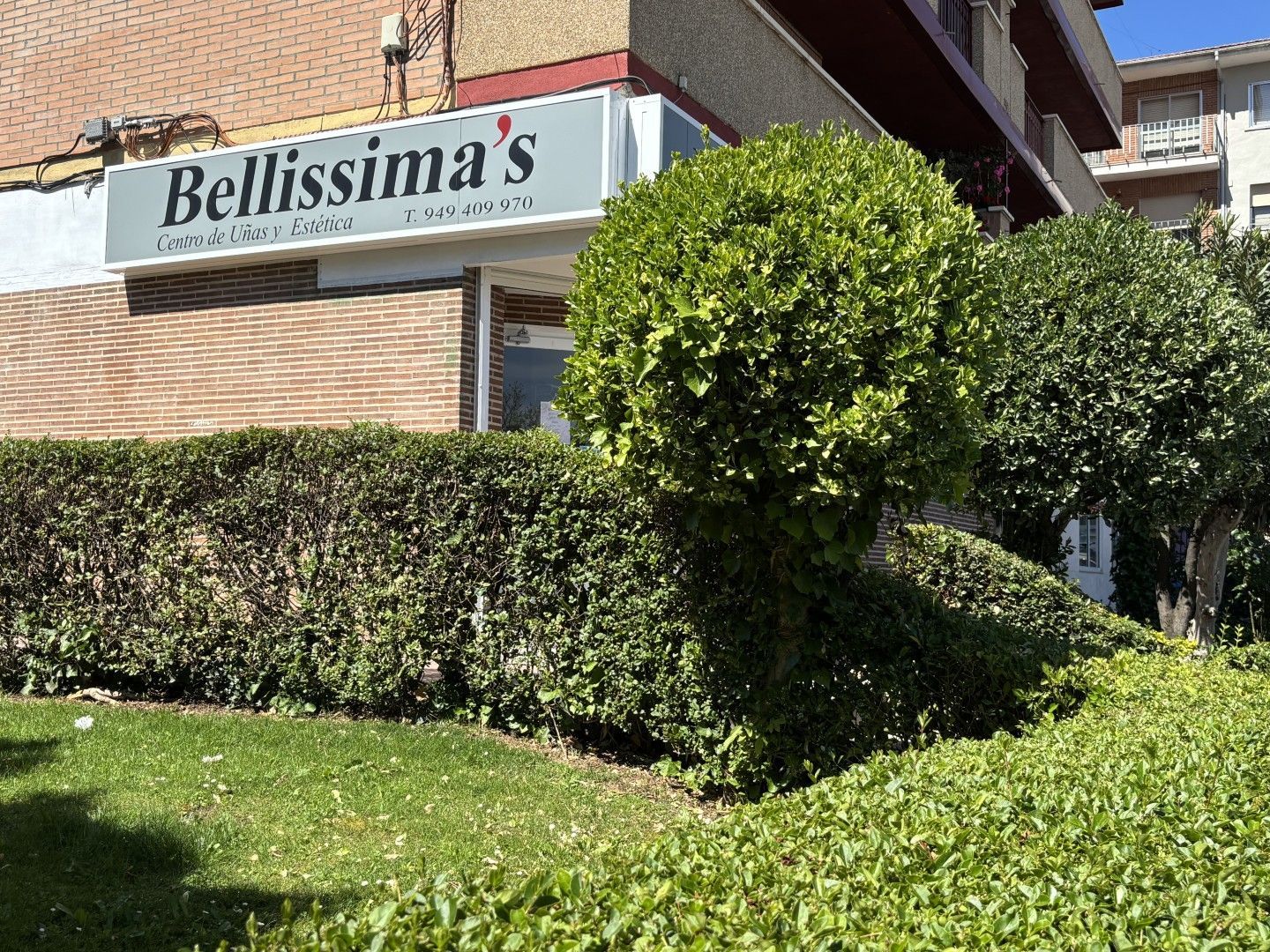 El letrero de la tienda Bellissima en un edificio de ladrillo, parcialmente oculto por setos y árboles bien cuidados.
