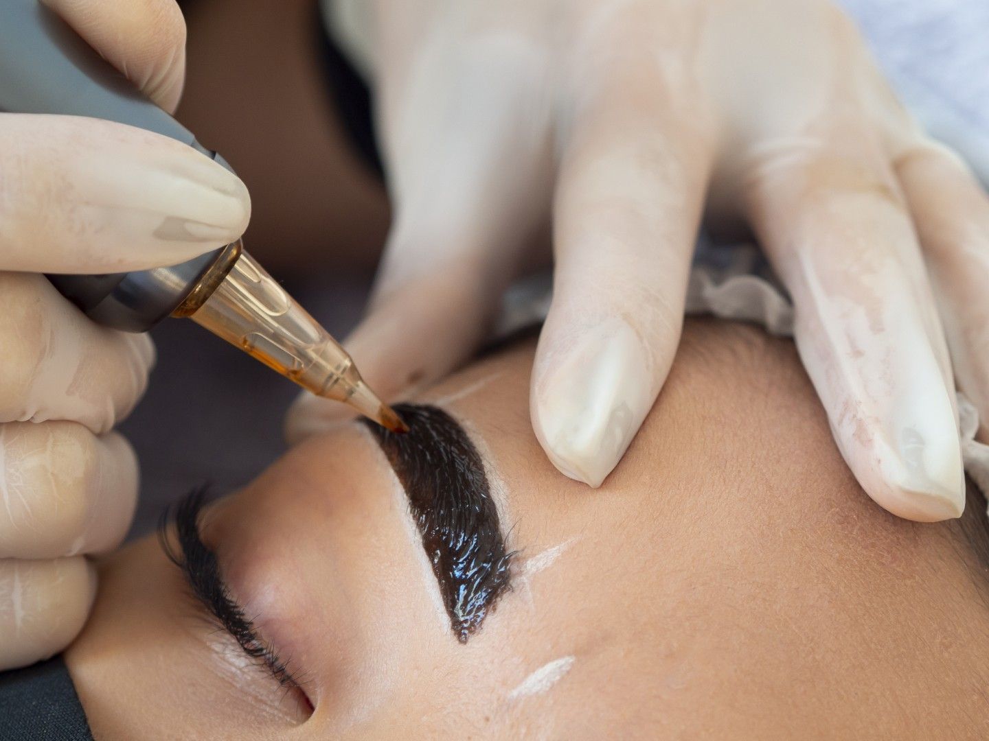 Manos enguantadas aplicando pigmento negro para cejas con un pincel durante un tatuaje cosmético.
