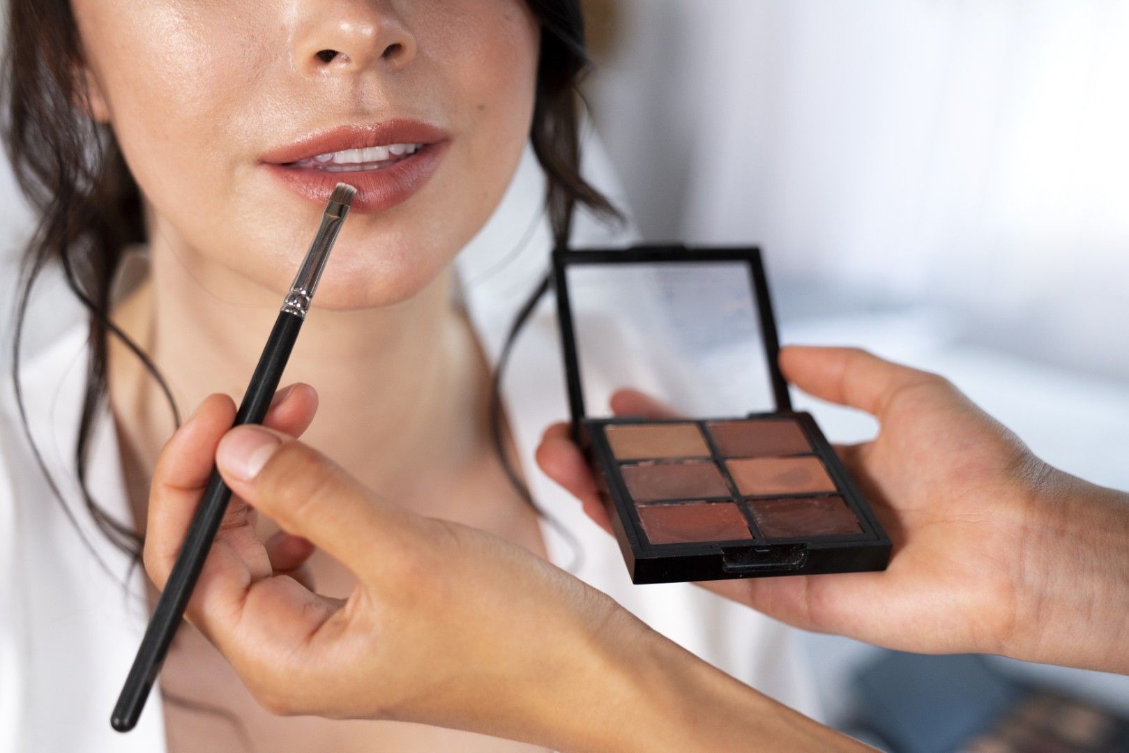 Una maquilladora aplica color de labios a una mujer mientras sostiene una paleta de sombras de ojos compacta.