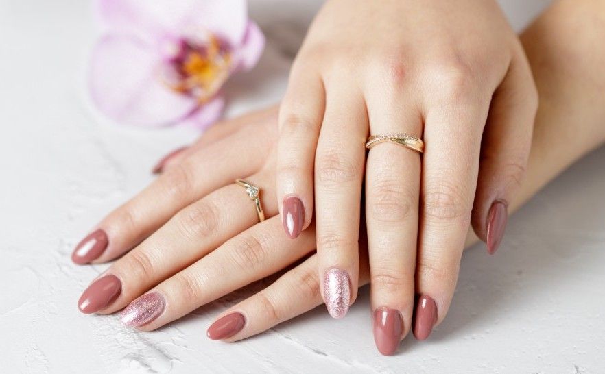 Manos bien cuidadas con uñas color nude rosado y anillos de oro descansando sobre una superficie blanca cerca de una flor.
