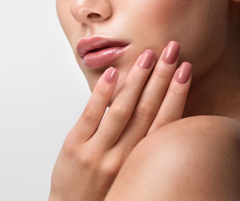 Primer plano del rostro y la mano de una persona con uñas rosas bien cuidadas sobre un fondo claro.