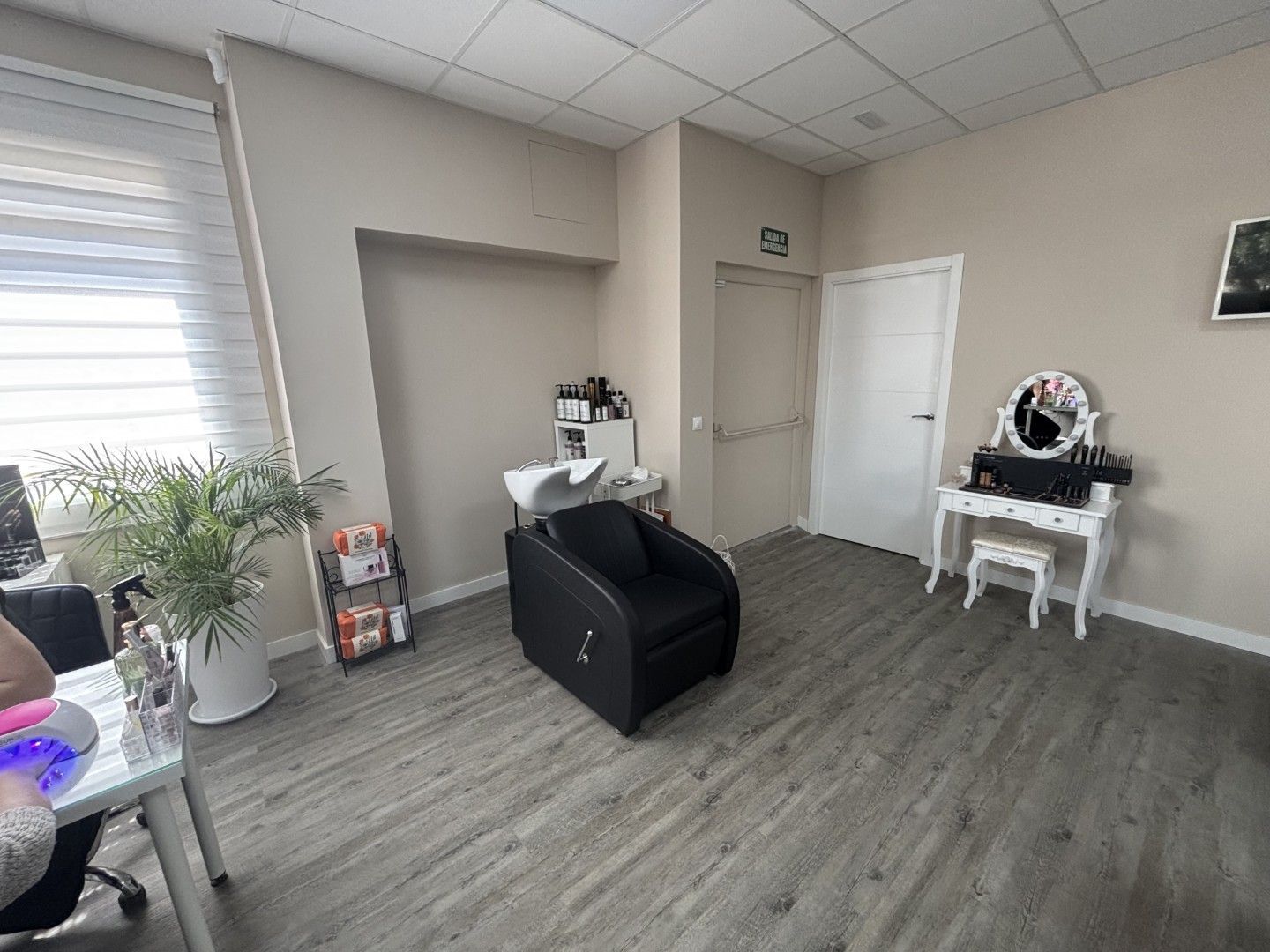 Sala de espera vacía con suelo gris, silla de peluquería negra, tocador blanco y plantas cerca de una ventana.