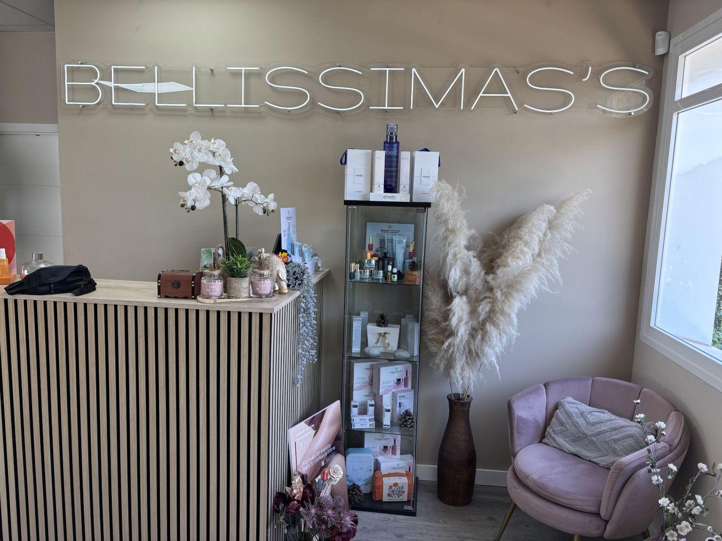Recepción de estilo boutique con letrero de neón, estante de productos, plantas y una silla malva en una habitación.
