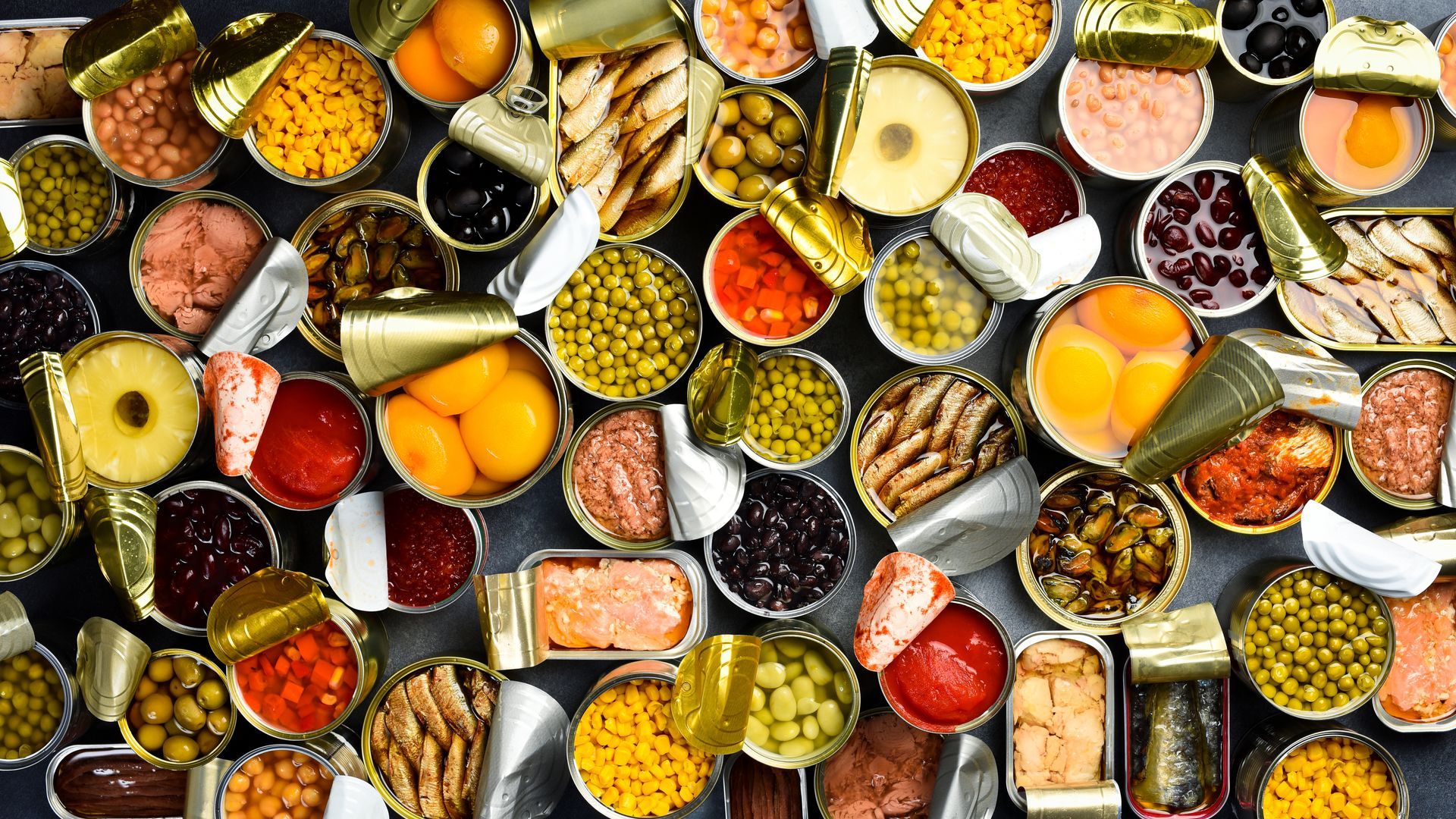 Vue de dessus de nombreuses boîtes de conserve ouvertes remplies de divers aliments, notamment des fruits, des légumes, du poisson et des sauces.