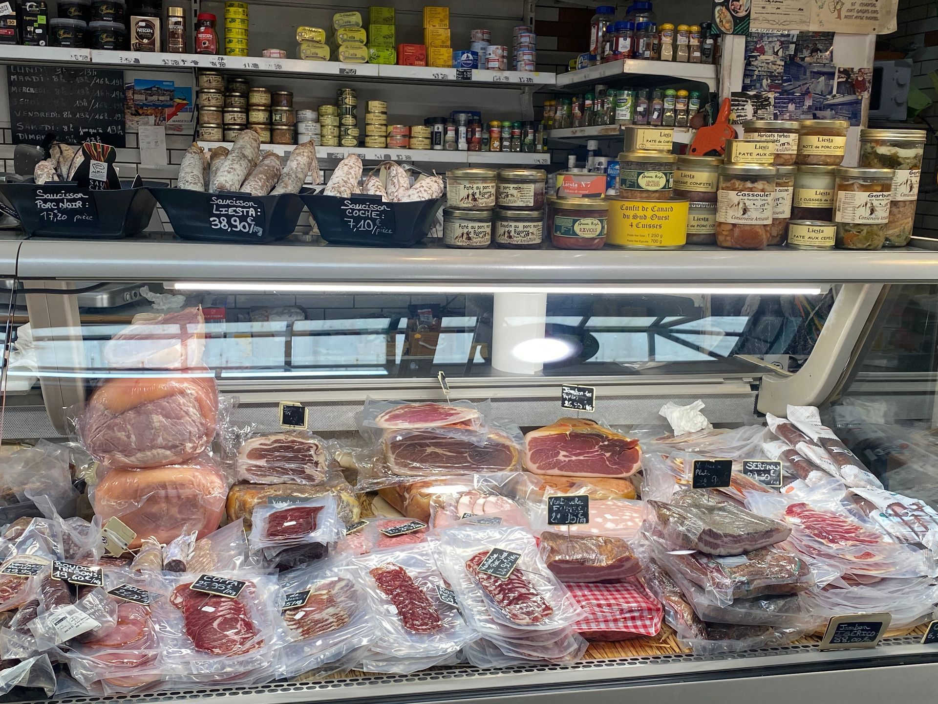 Un comptoir de charcuterie présentant diverses viandes et fromages dans des emballages transparents, exposés sous des étagères de bocaux et de petits produits.