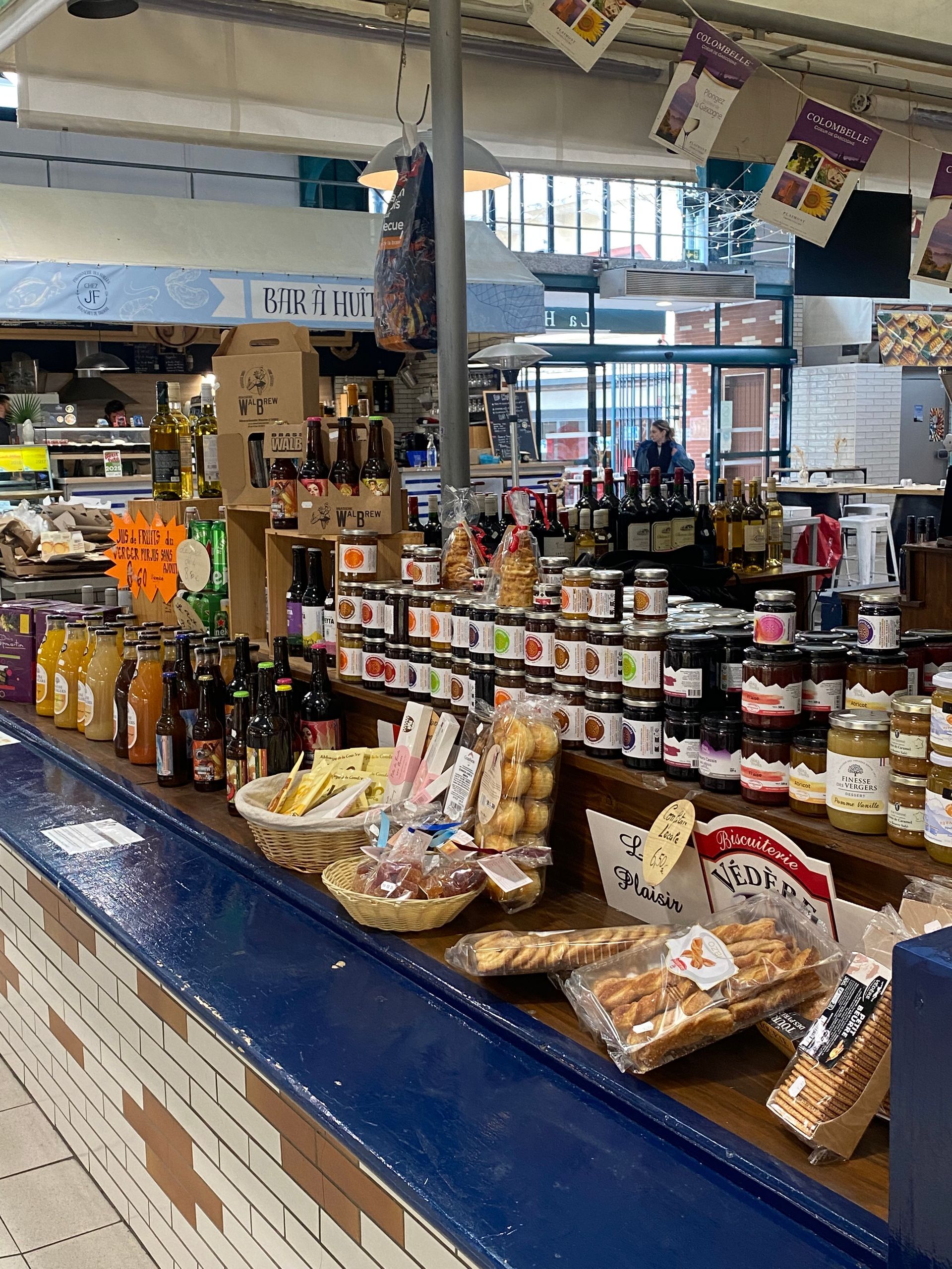 Dans un marché couvert, un comptoir bleu expose divers bocaux, des boissons en bouteille et des paniers de produits de boulangerie emballés.