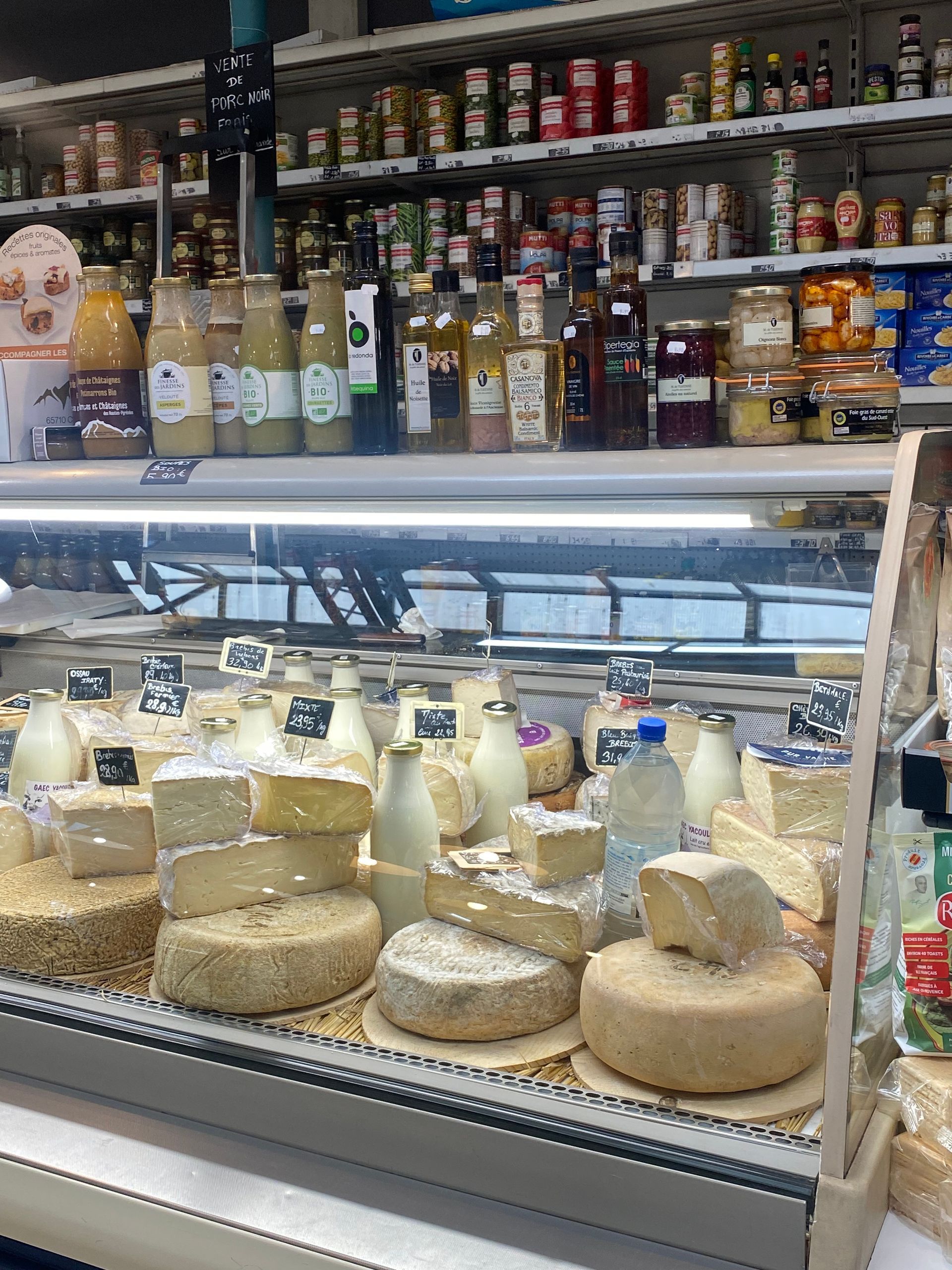 Une vitrine remplie de fromages ronds variés dans un magasin, avec des bouteilles et des bocaux sur des étagères en arrière-plan.