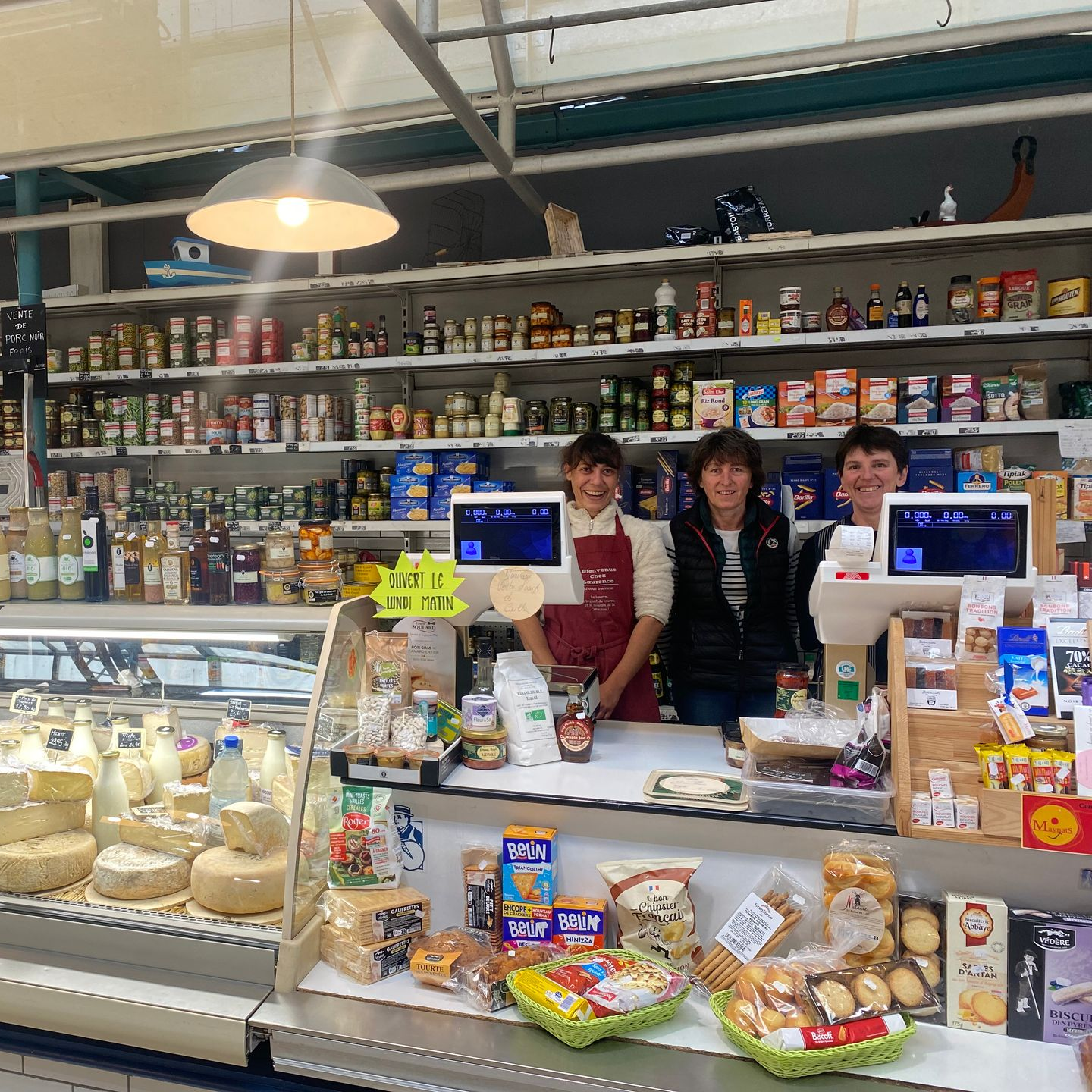 Trois employés sourient derrière un comptoir de charcuterie garni de fromages, de pain et d'étagères de produits emballés.